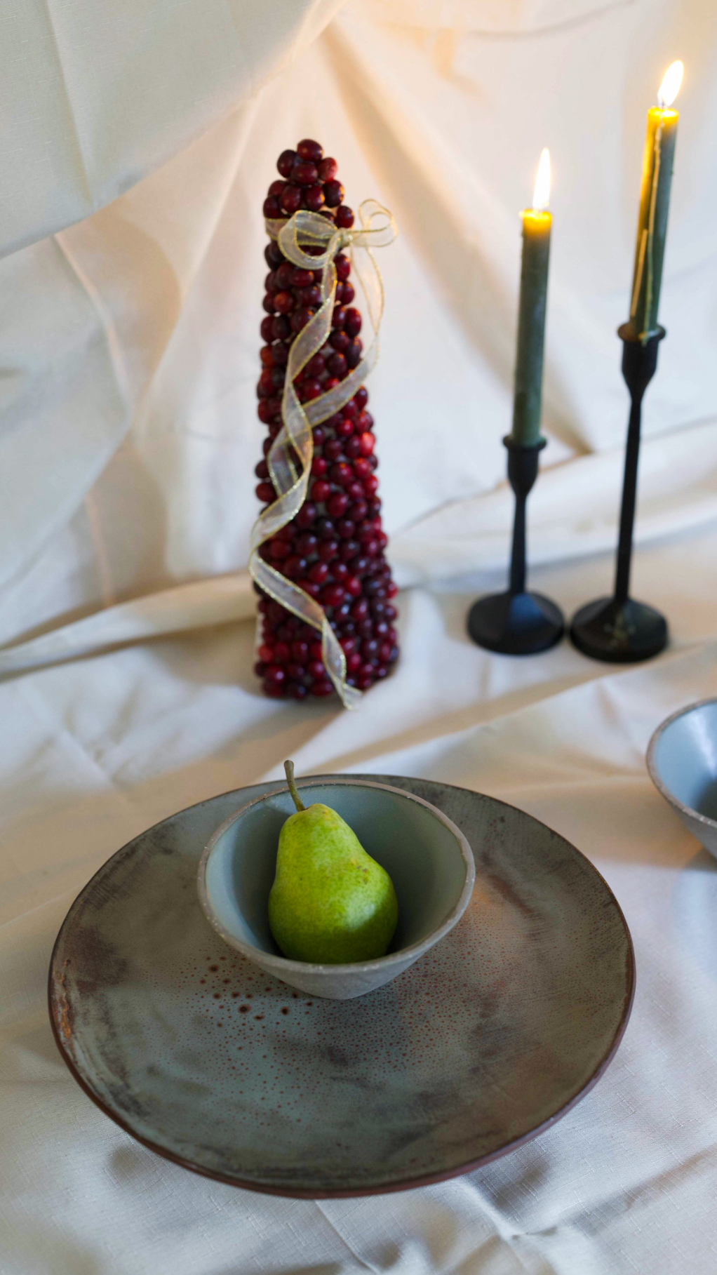 Decorative setup with a green pear in a bowl, cranberry and ribbon arrangement, and candles on a white surface.